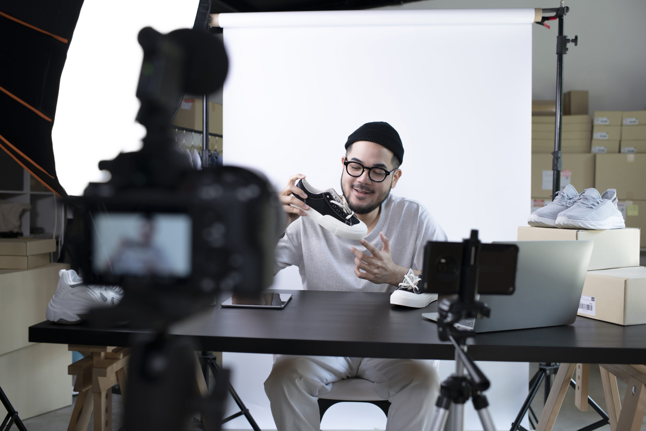 A hip, well-dressed male influencer with a shoe in his hand sits on a chair in front of a white screen and is filmed.