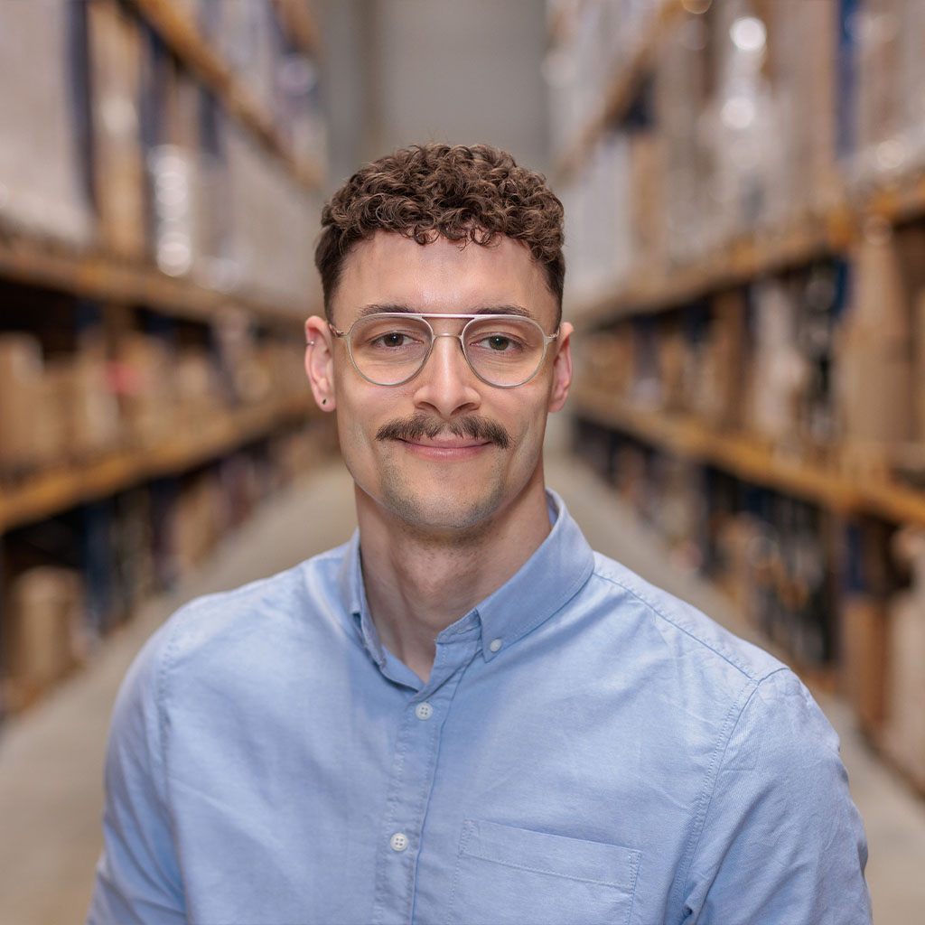 A male person is standing between two shelves. The person is wearing a blue shirt and glasses. 